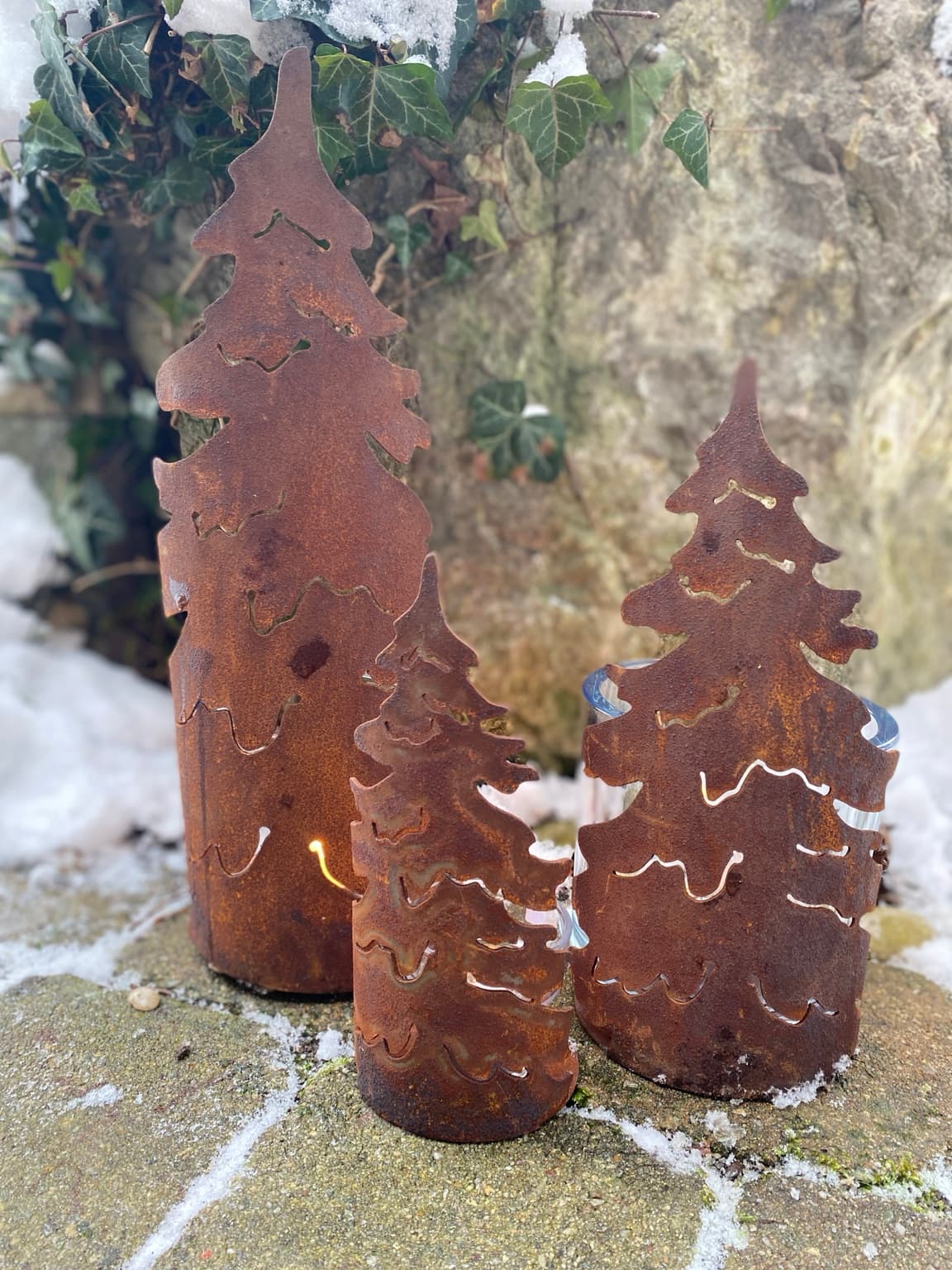 Beleuchtete Edelrost-Tannen im Schnee vor Efeu und Naturstein